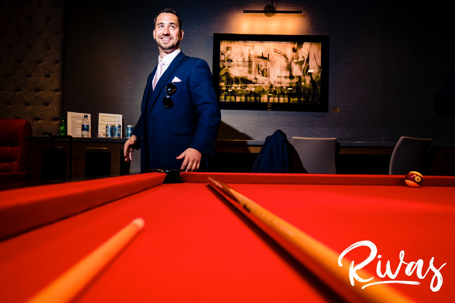 A bright, candid portrait taken across a pool table of a groom standing at the edge, laughing at his groomsmen on the day of his wedding. 