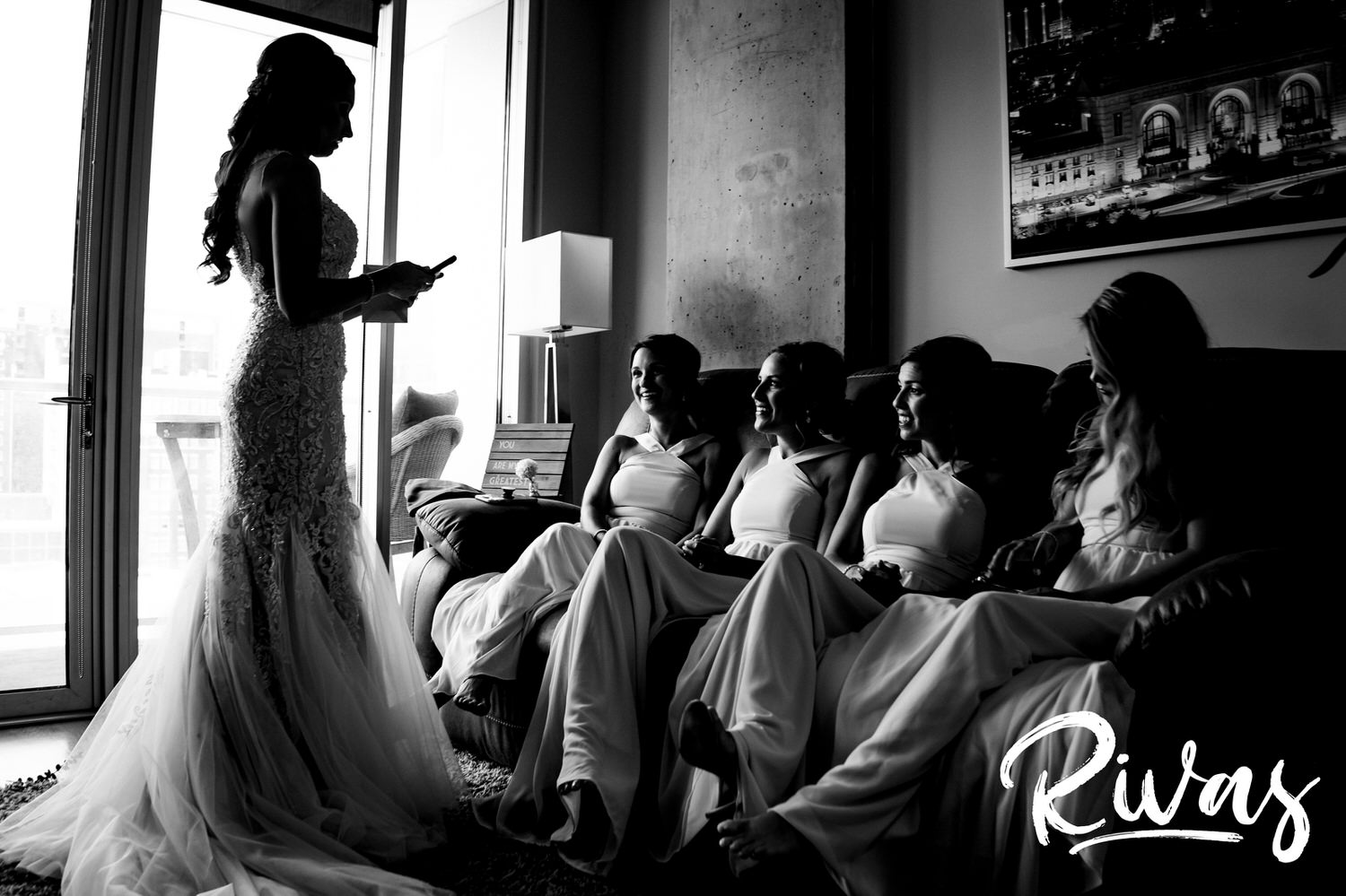A candid black and white picture of a bride holding an envelope, looking down at a group of bridesmaids sitting on a couch on the morning of her wedding in downtown Kansas City. 