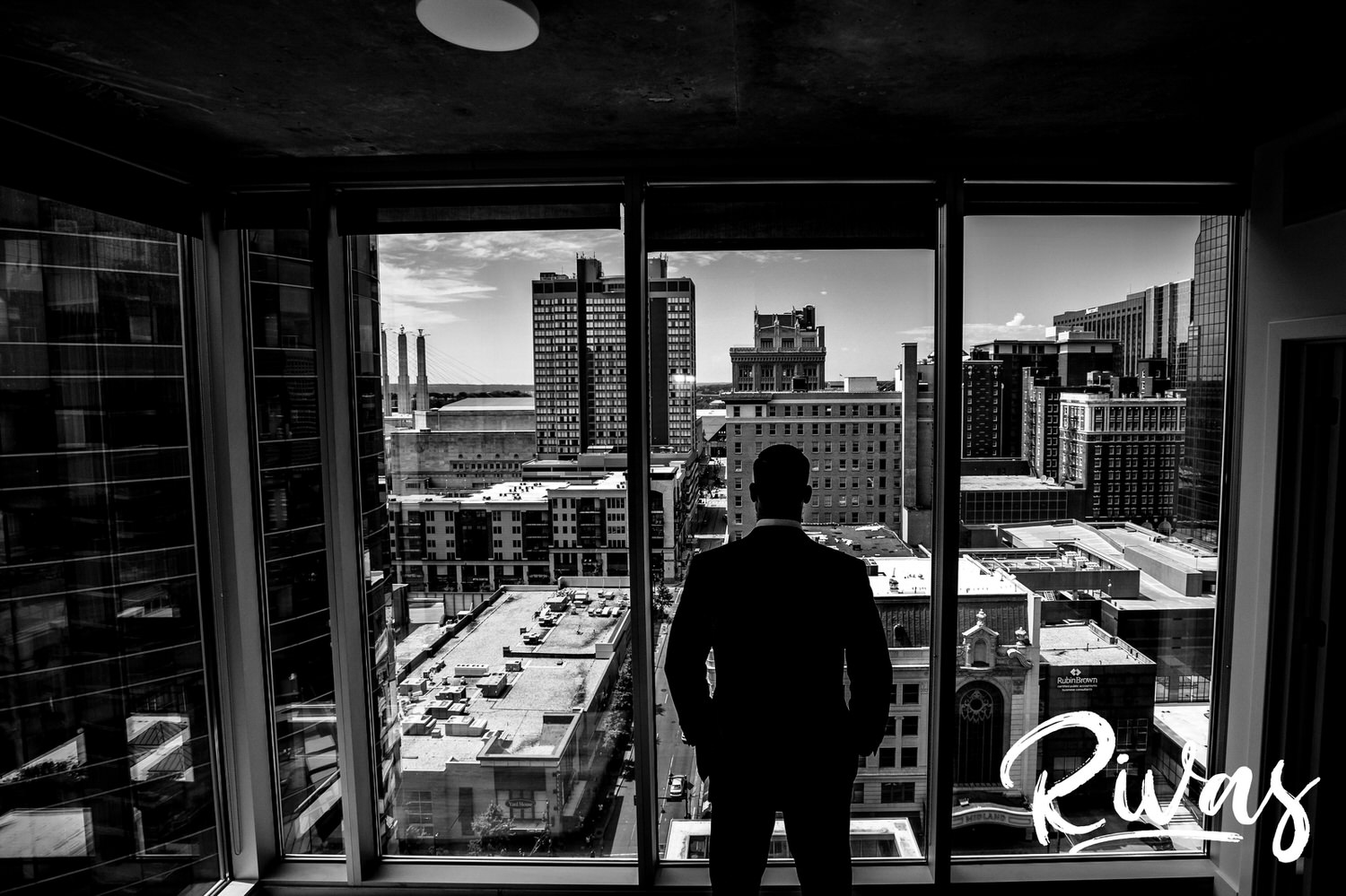 A candid, black and white picture taken from behind as a groom stands with his hands in his pocket in front of a giant window, looking out over Downtown Kansas City on his wedding day. 