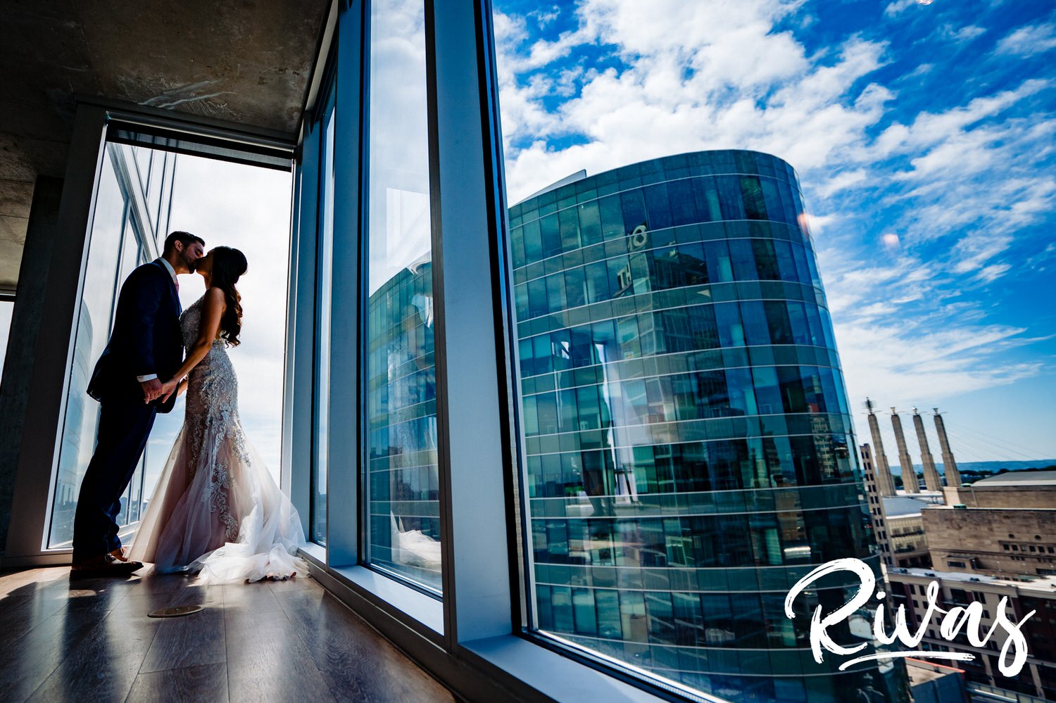 A vibrant portrait of a bride and groom holding hands and sharing a kiss in front of floor to ceiling windows overlooking downtown Kansas City on their wedding day. 
