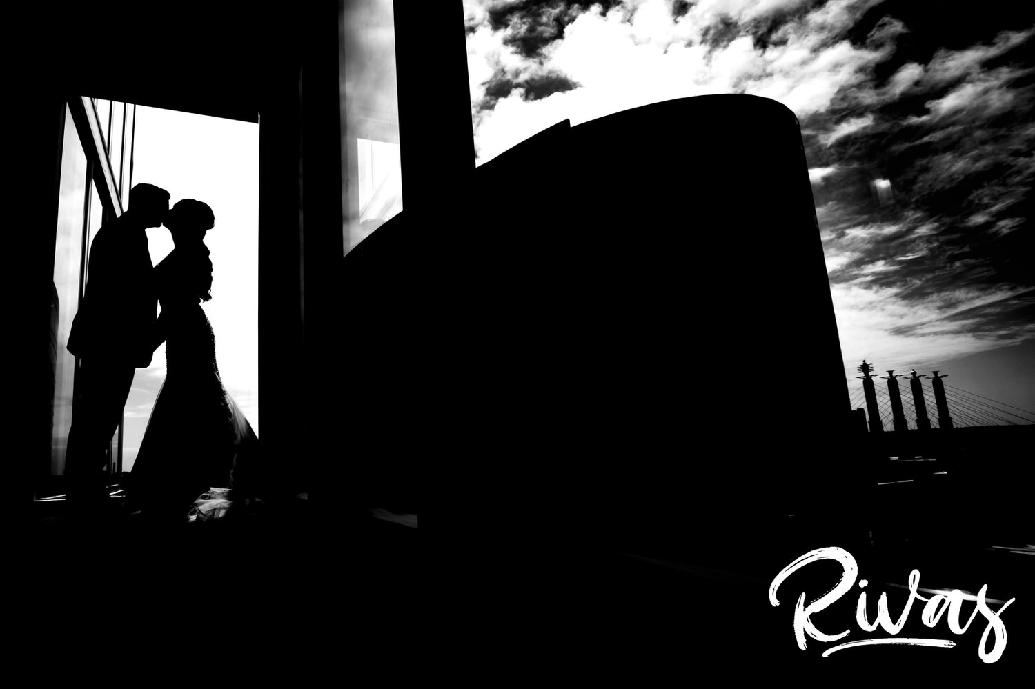 A dramatic, black and white portrait of a bride and groom leaning in for a kiss, silhouetted against the skyline in Kansas City on their wedding day. 