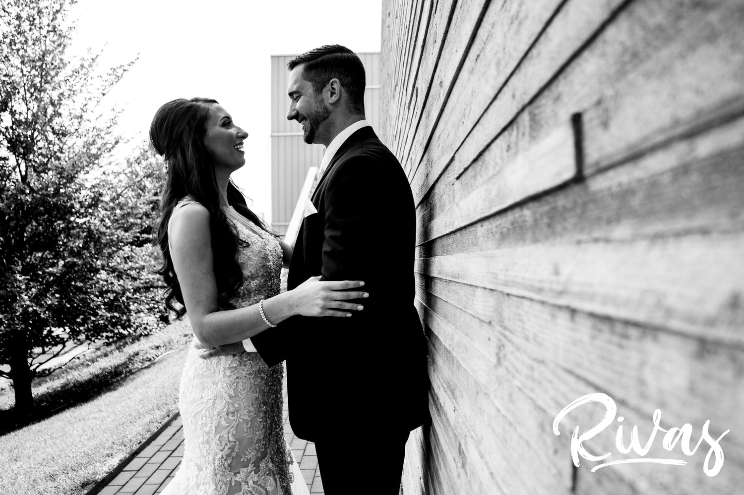 A casual, black and white picture of a bride and groom leaning up against a gray stone wall, sharing an embrace and laughing together on their wedding day. 