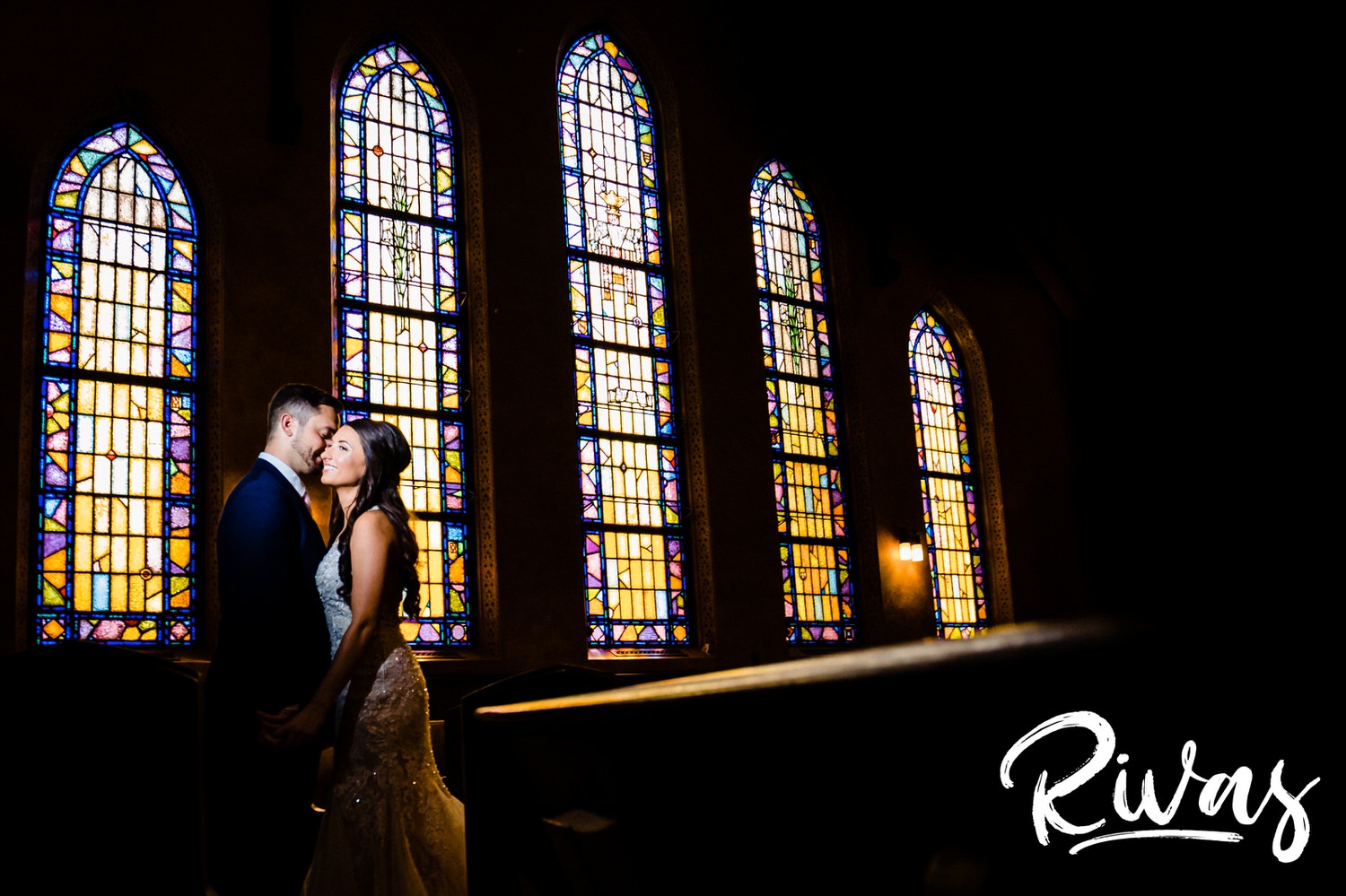 A bright, dramatic portrait of a bride and groom sharing a kiss in front of yellow stained glass windows on their wedding day.