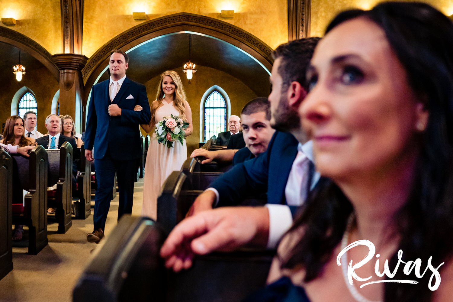 A candid picture, taken from just behind the mother of the bride, of a bridesmaid and groomsman walking down the aisle as the mom looks back at the groom. 