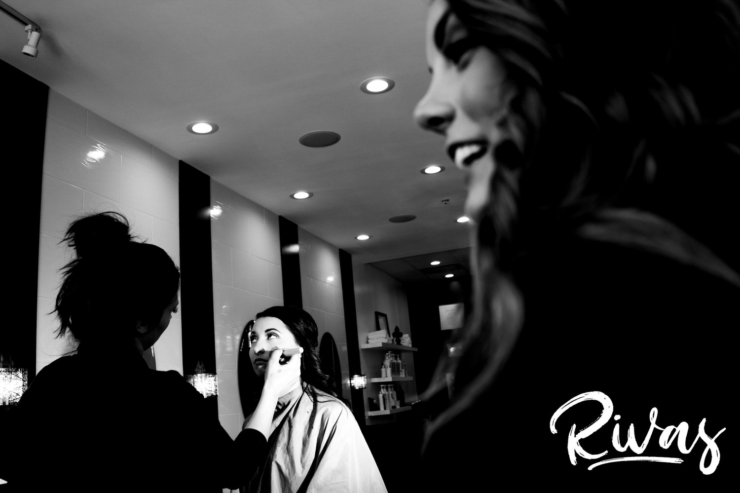 A candid black and white picture of a bride getting her makeup done as a bridesmaid laughs in the foreground on the morning of her wedding in downtown Kansas City. 