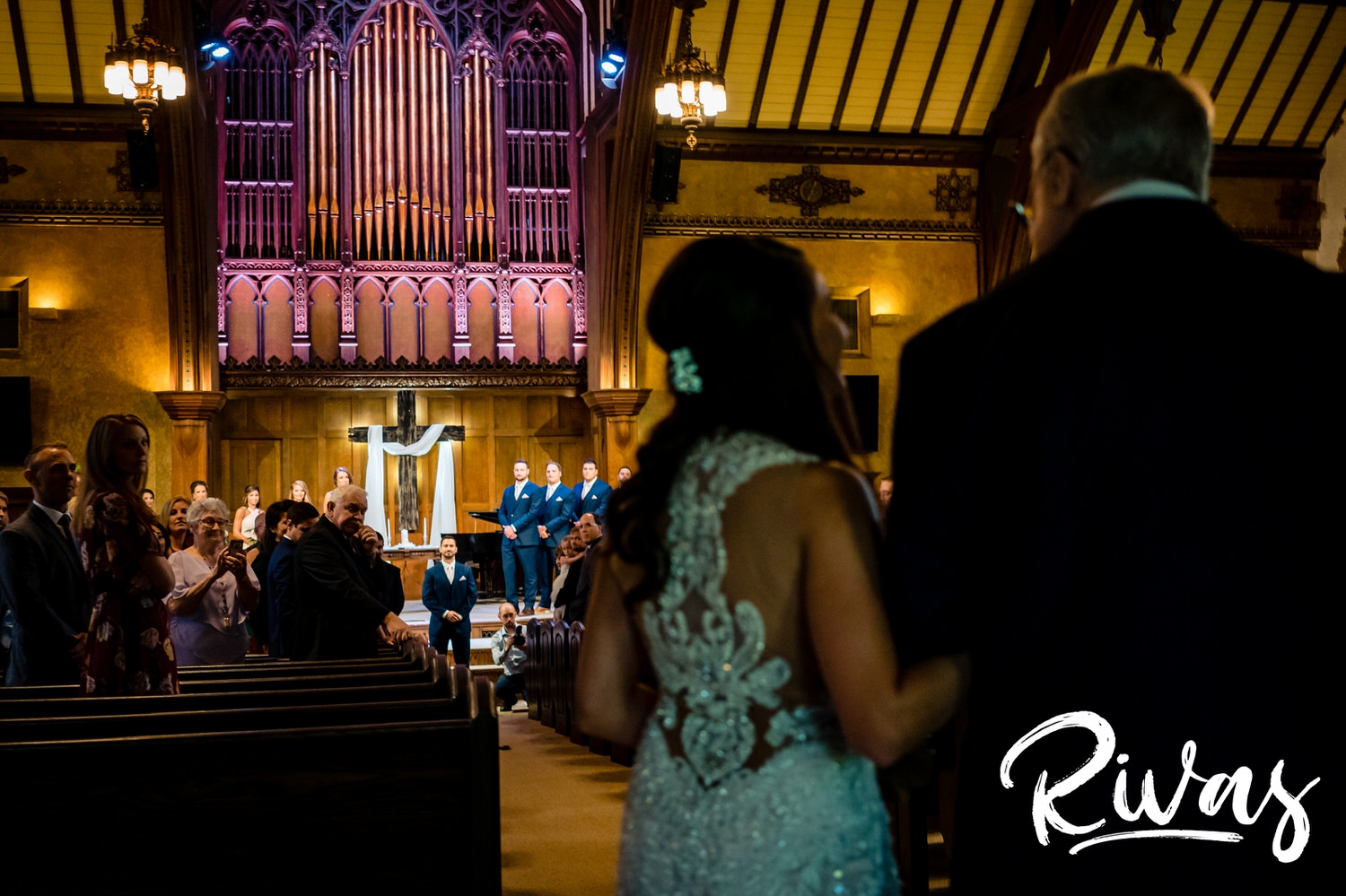 A candid, wide, picture taken from the back of the church as a bride and her grandpa turn to walk down the main aisle of the church, as her groom and the rest of the wedding party is visible in the background of the image. 