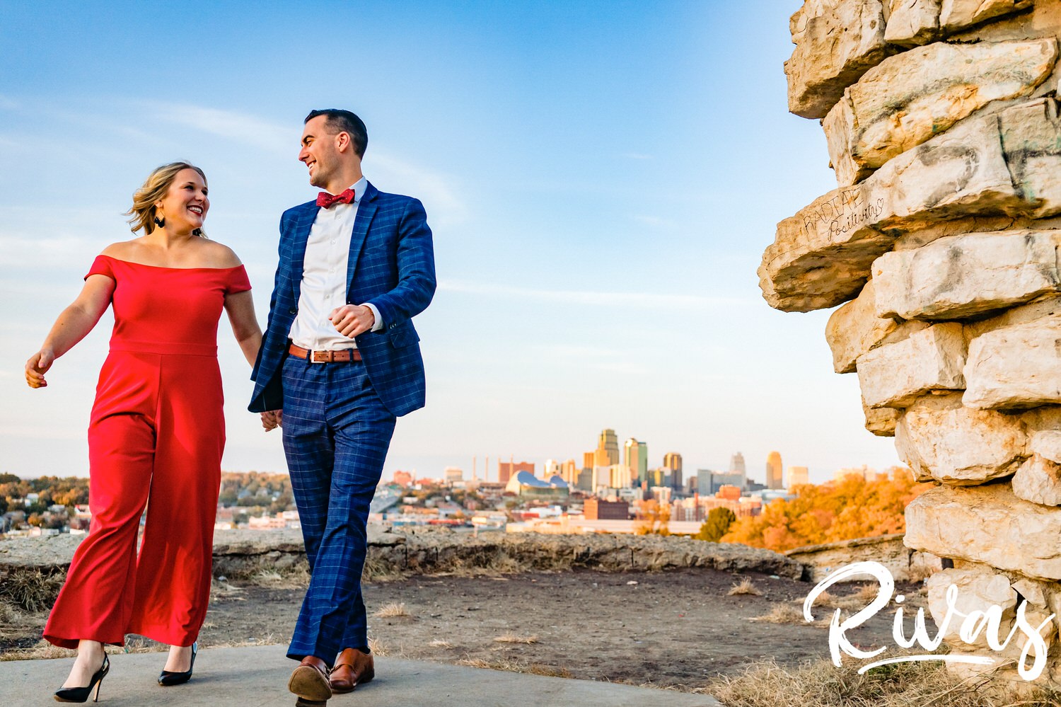 A bright, vibrant picture of a woman in a red jumpsuit and a man in a blue suit holding hands and walking towards the camera with the Kansas City skyline in the background during their engagement session at The Scout. 