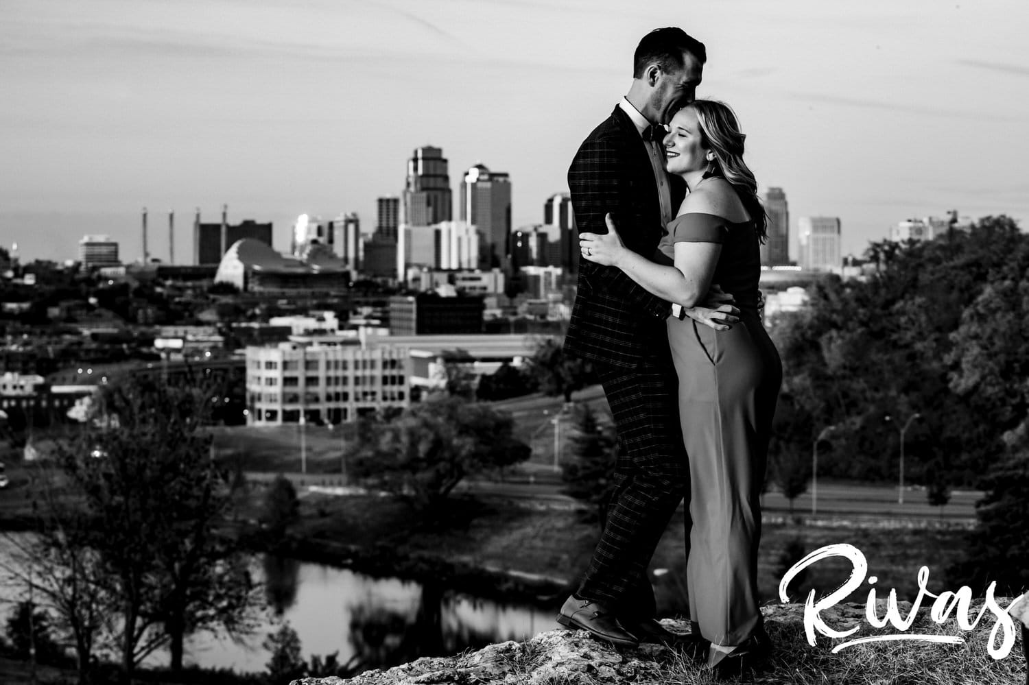 A candid black and white picture of a woman looking adoringly up at her fiance as they stand in front of the Kansas City Skyline at The Scout during their engagement session in Kansas City. 