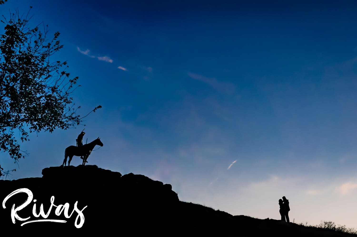 A very wide, silhouette picture of an engaged couple sharing an embrace as they stand just downhill from The Scout statue in Kansas City. 