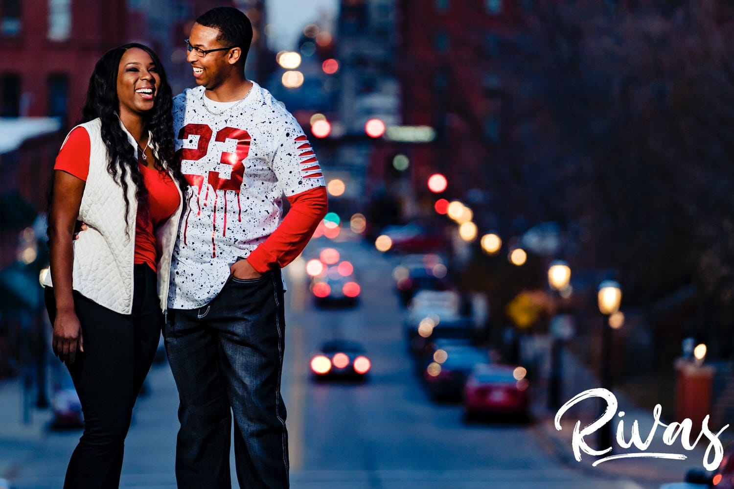 A bright, candid picture of an engaged couple standing on top of a hill, sharing an embrace and laughing together with the city streets and lights of downtown Kansas City visible in the background. 