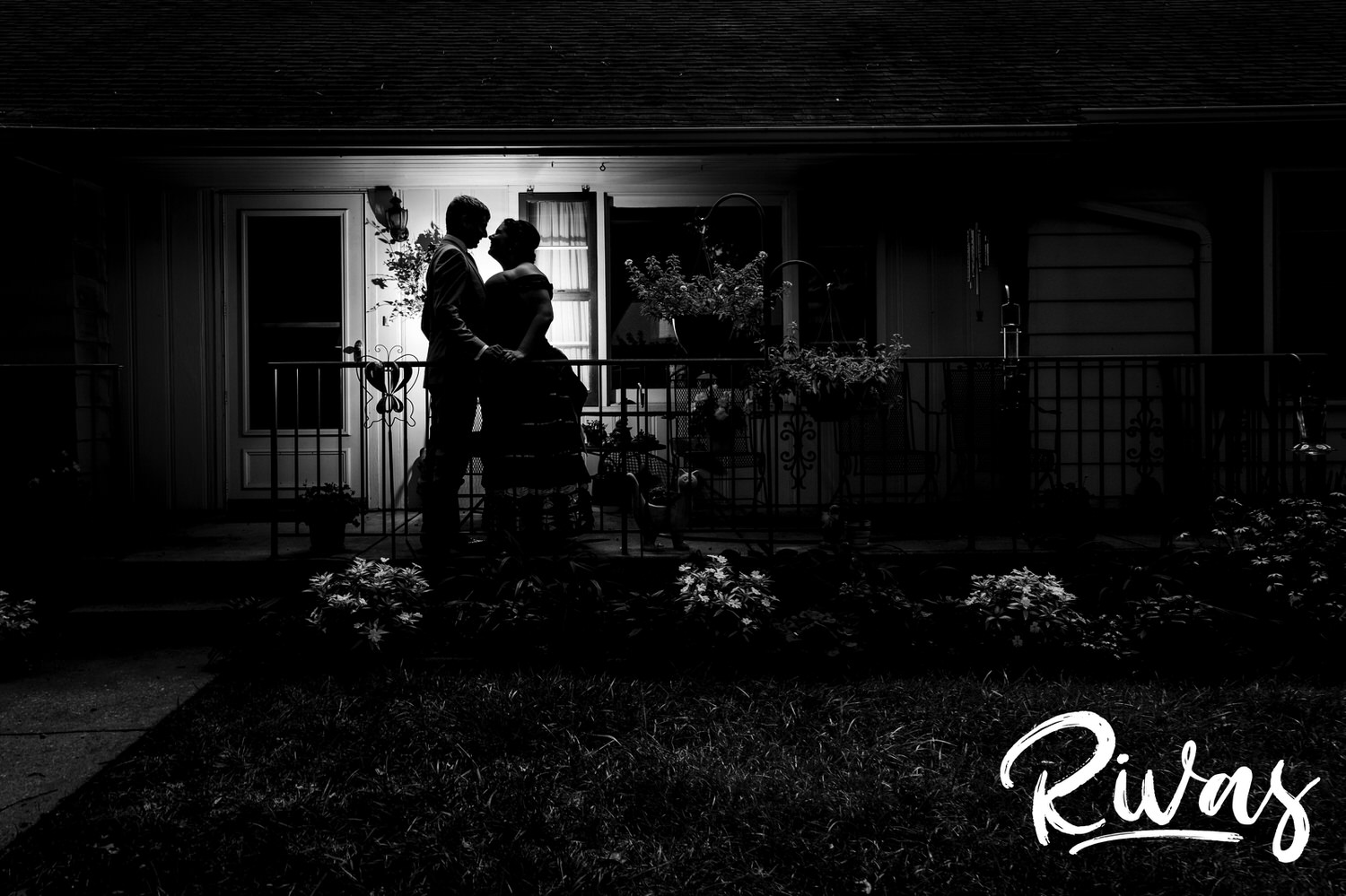 A black and white portrait of a bride and groom sharing an embrace as they stand on the front porch of a family home on their intimate backyard summer wedding day. 