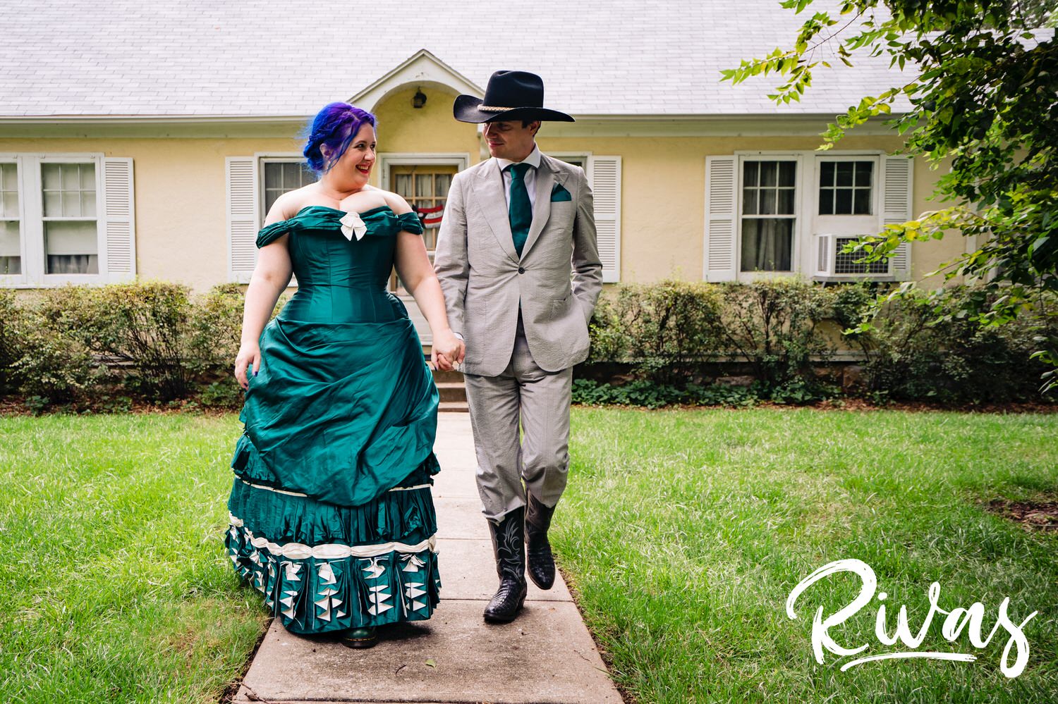 A candid picture of a bride in a custom jade gown and a groom in a tux with cowboy boots and hat holding hands and walking down the sidewalk of a family home on their summer wedding day in Kansas City. 