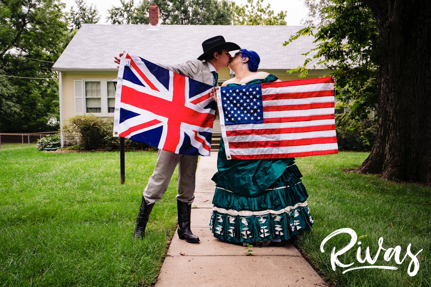 A candid picture of a bride in a custom jade wedding gown holding an American flag sharing a kiss with a groom wearing a cowboy hat and boots holding a British flag on their wedding day in Kansas City. 