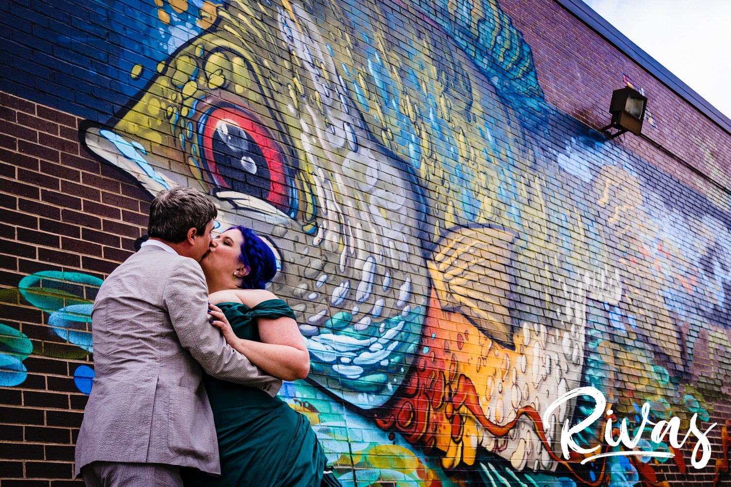 A candid picture of a bride in a jade wedding gown and a groom in cowboy boots sharing a kiss as they stand in front of a giant, colorful fish mural in downtown Overland Park. 