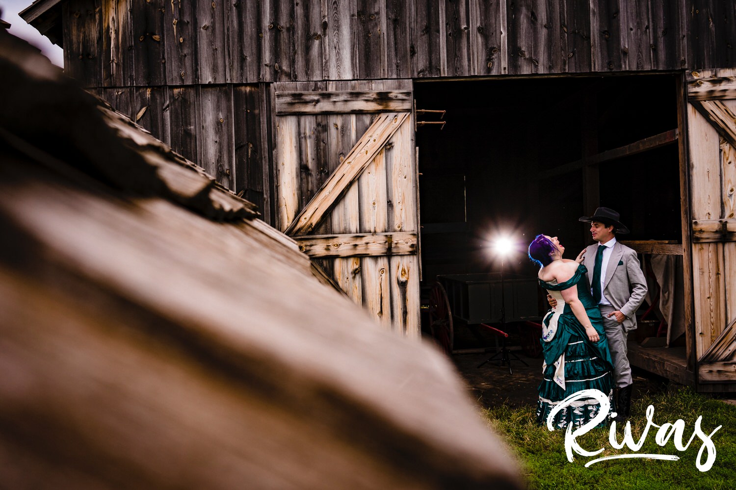 A candid portrait of a bride and groom laughing together as they share an embrace in front of a dark wood barn at Mahaffie Stage Coach Stop on their summer wedding day in Kansas City. 