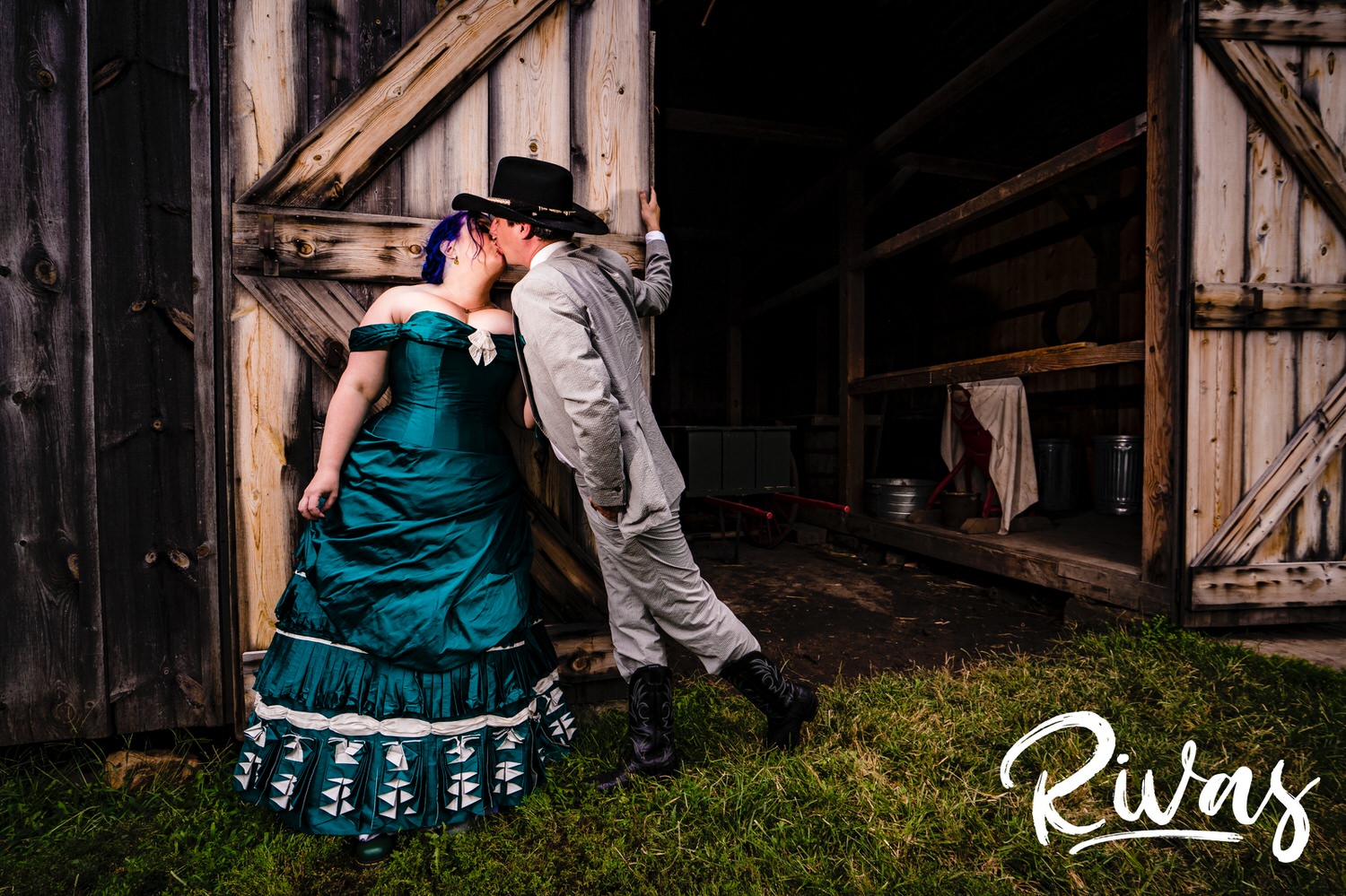 A candid picture of a groom in a cowboy hat and boots leaning in to give his bride in a jade gown a kiss as she stands in front of a barn's open door at Mahaffie Stage Coach Stop on their summer wedding day. 