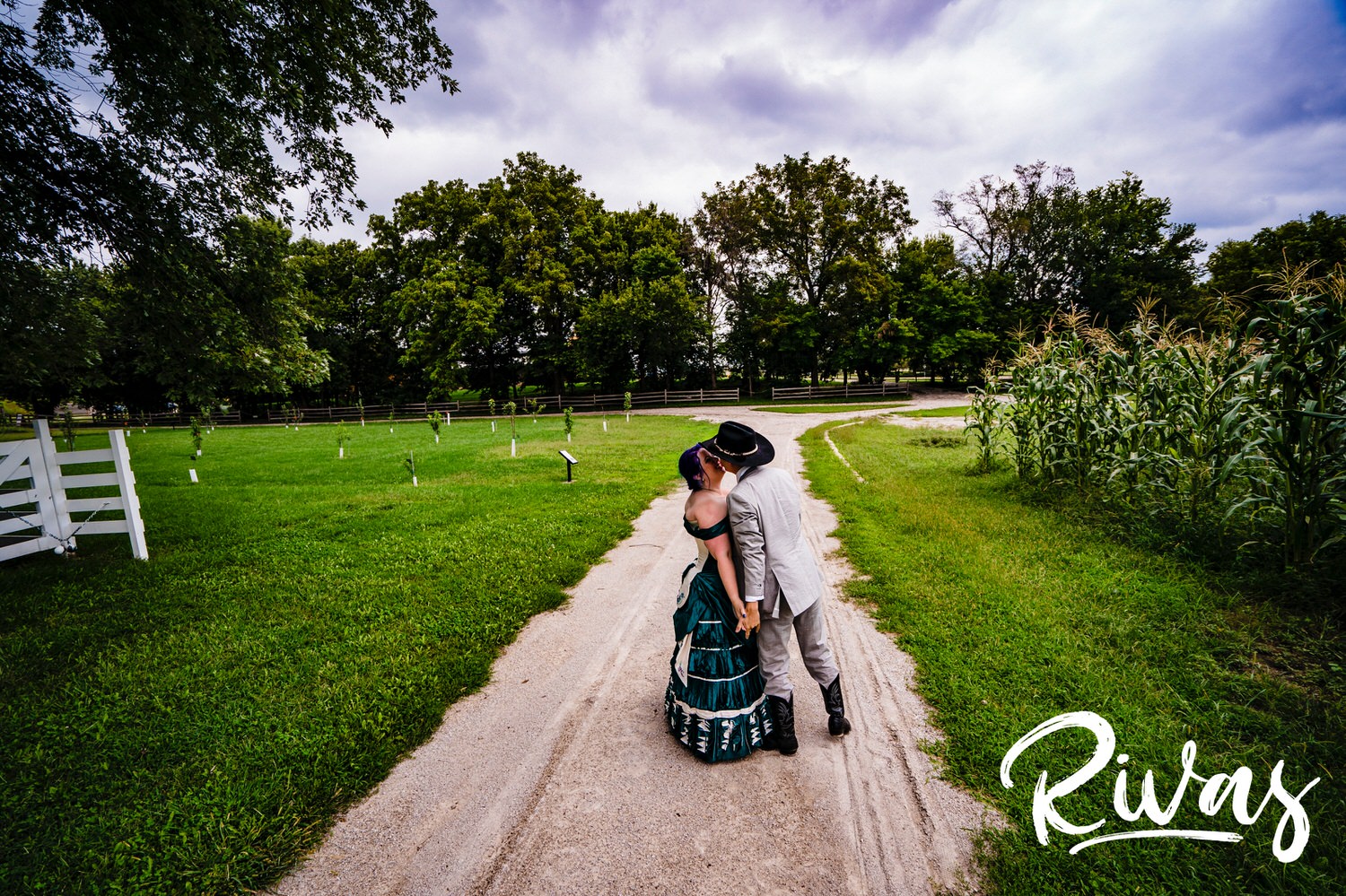 A candid picture of a bride in a jade gown and a groom in cowboy hat and boots holding hands and walking down a gravel road with barns visible in the background on their intimate summer wedding day in Kansas City. 