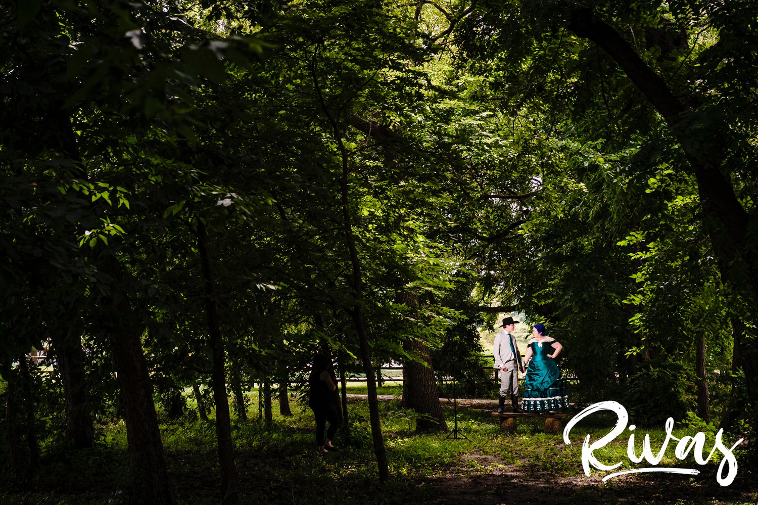 A wide picture of a bride and groom standing under a canopy of green tree leaves at Mahaffie Stagecoach Stop on their intimate summer wedding day in Kansas City. 
