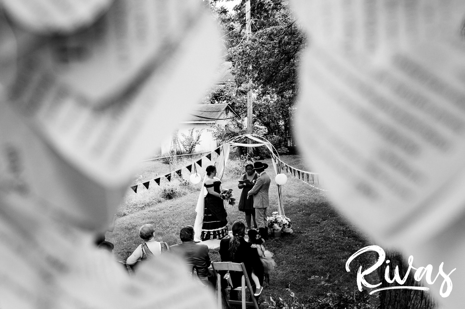 A candid black and white picture taken during a backyard wedding ceremony of a bride and groom standing under an arbor during a reading during their wedding ceremony. 
