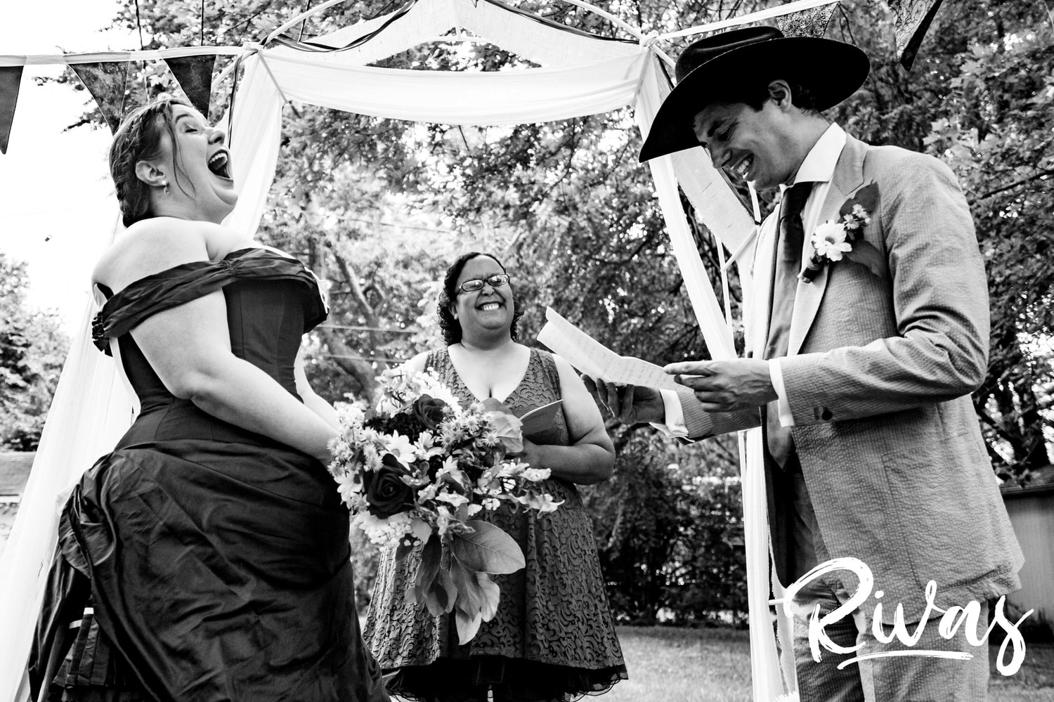 A candid black and white picture of a groom in a cowboy hat reading his Country-Song-inspired vows to his bride, who is laughing hysterically during their intimate backyard summer wedding ceremony. 