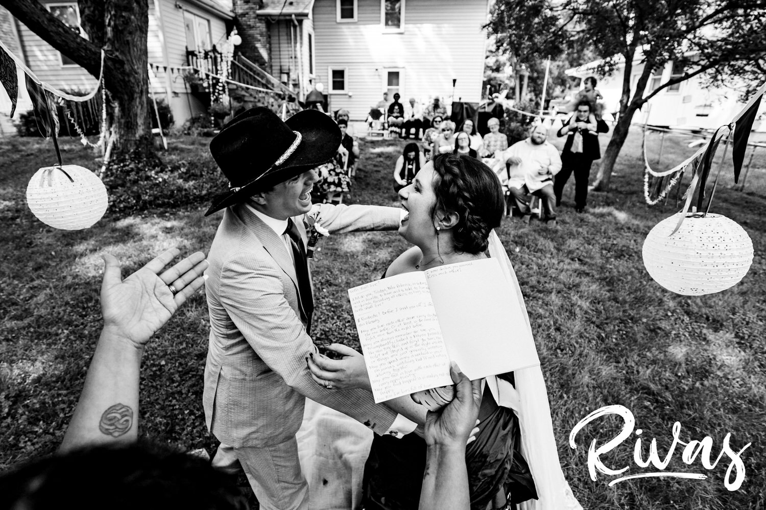 A candid black and white picture of an officiate encouraging a bride and groom to share a kiss at the end of their backyard wedding ceremony in Kansas City. 