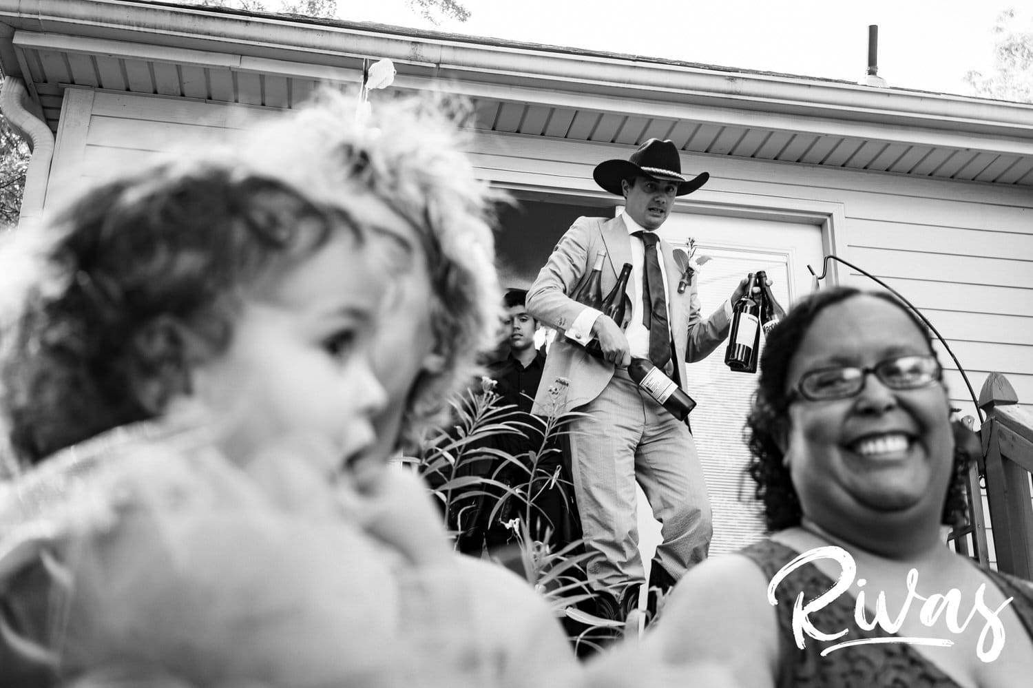 A candid black and white picture with a woman laughing in the foreground and a groom in a cowboy hat holding 6 bottles of champagne walking down a set of porch steps during his backyard wedding reception. 