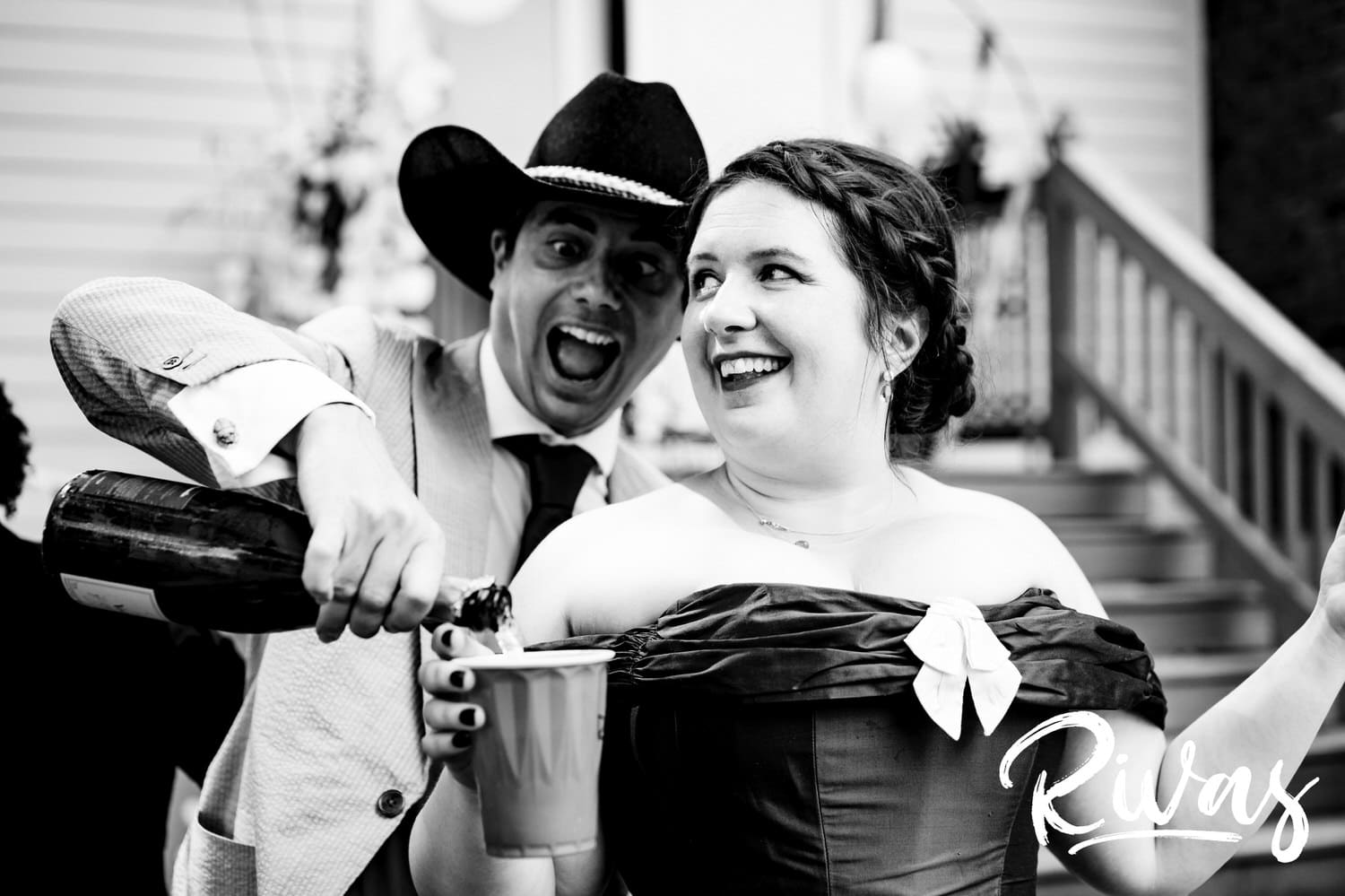 A candid black and white picture of a groom wearing a cowboy hat pouring champagne into his bride's red solo cup during their intimate backyard wedding reception in Kansas City. 