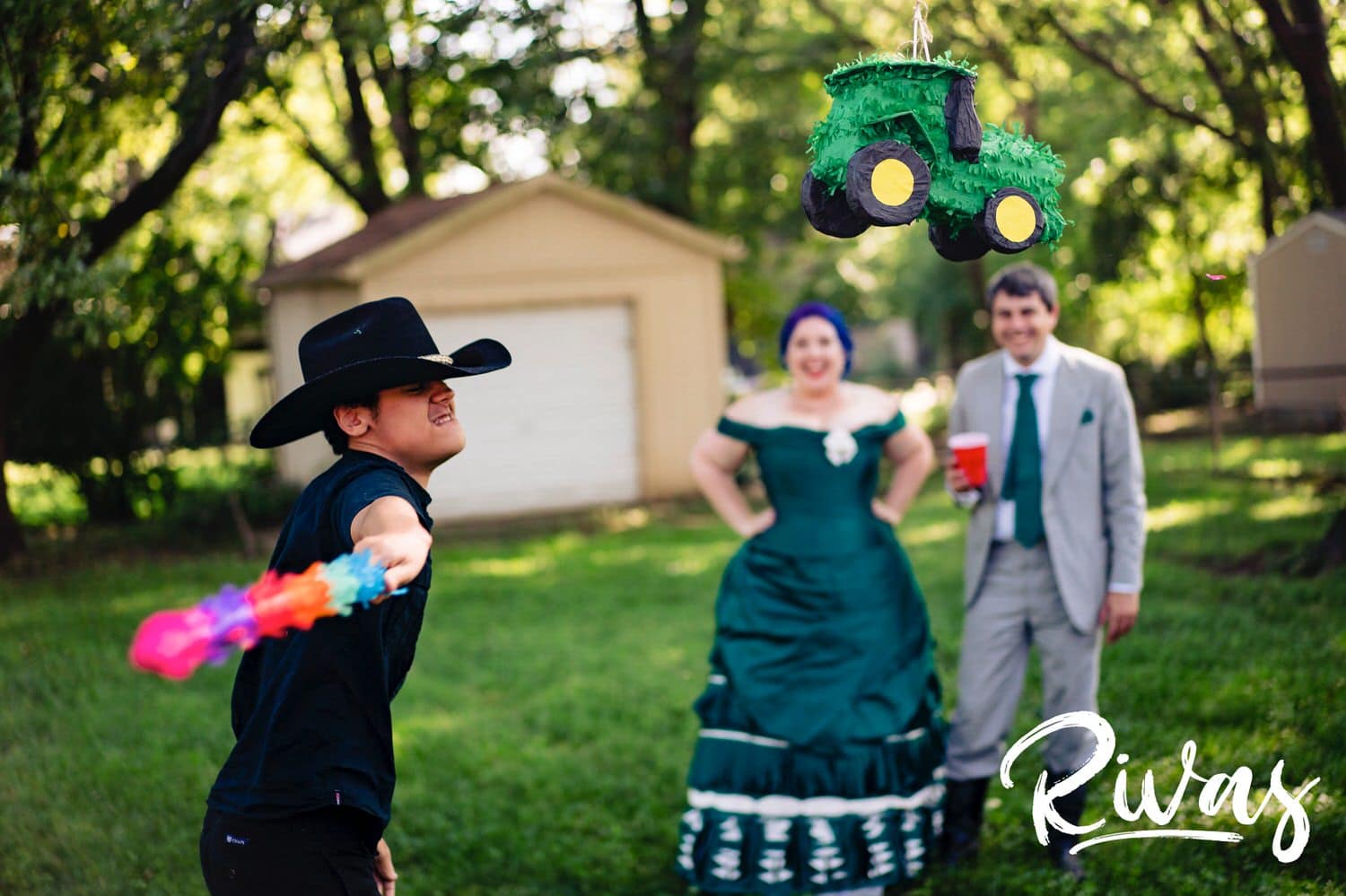 A candid picture of a teenager in a black cowboy hat wildly swinging a colorful stick at  a green tractor pinata while a bride and groom look on in the background with smiles on their faces during their intimate backyard summer wedding reception. 
