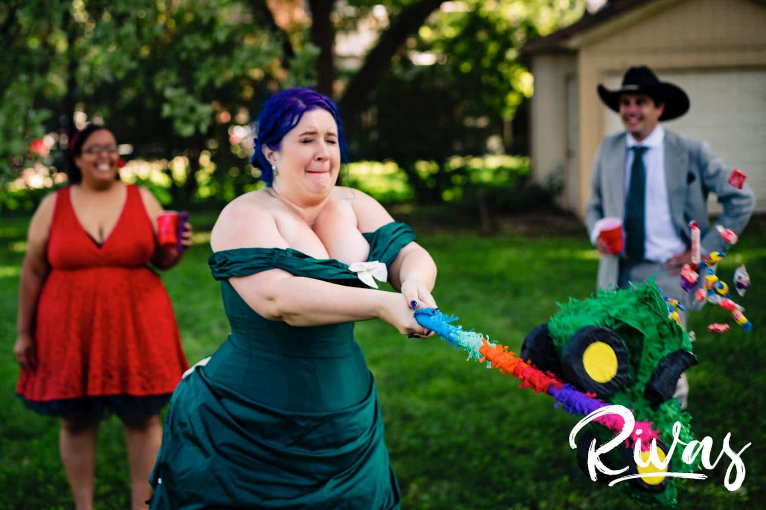 A candid picture of a bride in a jade gown hitting a green tractor pinata with a colorful stick as it explodes and candy spills out during her intimate backyard wedding reception in Kansas City. 
