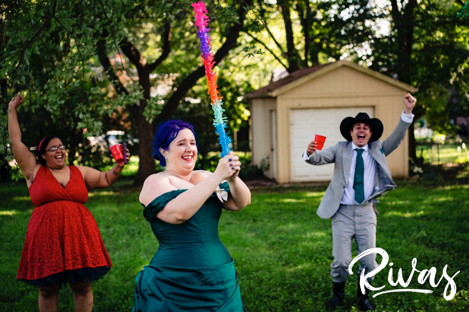 A candid picture of a bride in a jade wedding gown holding a colorful stick up in triump as her groom and friend cheer on behind her after she's blown-open a pinata during her intimate backyard wedding ceremony. 