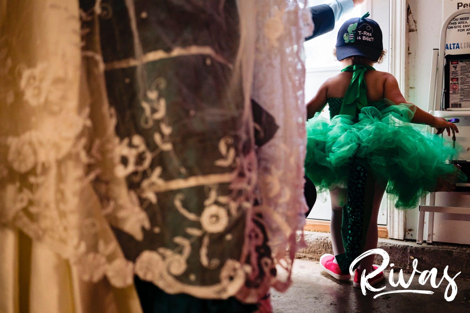 A candid picture taken from behind a bride wearing a jade gown and Belgian lace veil of a flower girl, wearing a bright green, sequined tutu with a sequined dinosaur tail and a "t-rex is best" backwards hat on, of the flower girl getting ready to walk down the wedding aisle. 