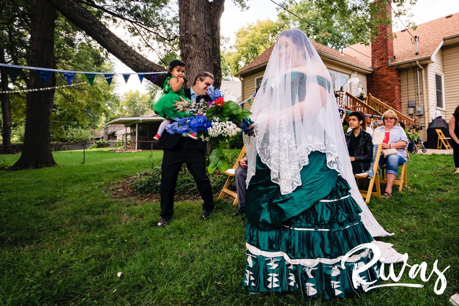 A candid picture of a bride in a custom jade gown and Belgian lace veil walking down the aisle during her intimate backyard wedding ceremony. 
