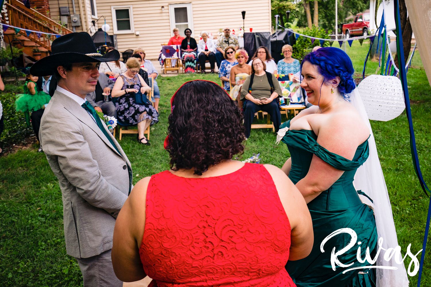 A candid picture taken from behind the officiate of a bride in a jade gown and a groom in a cowboy hat standing together during their intimate backyard wedding ceremony in Kansas City. 