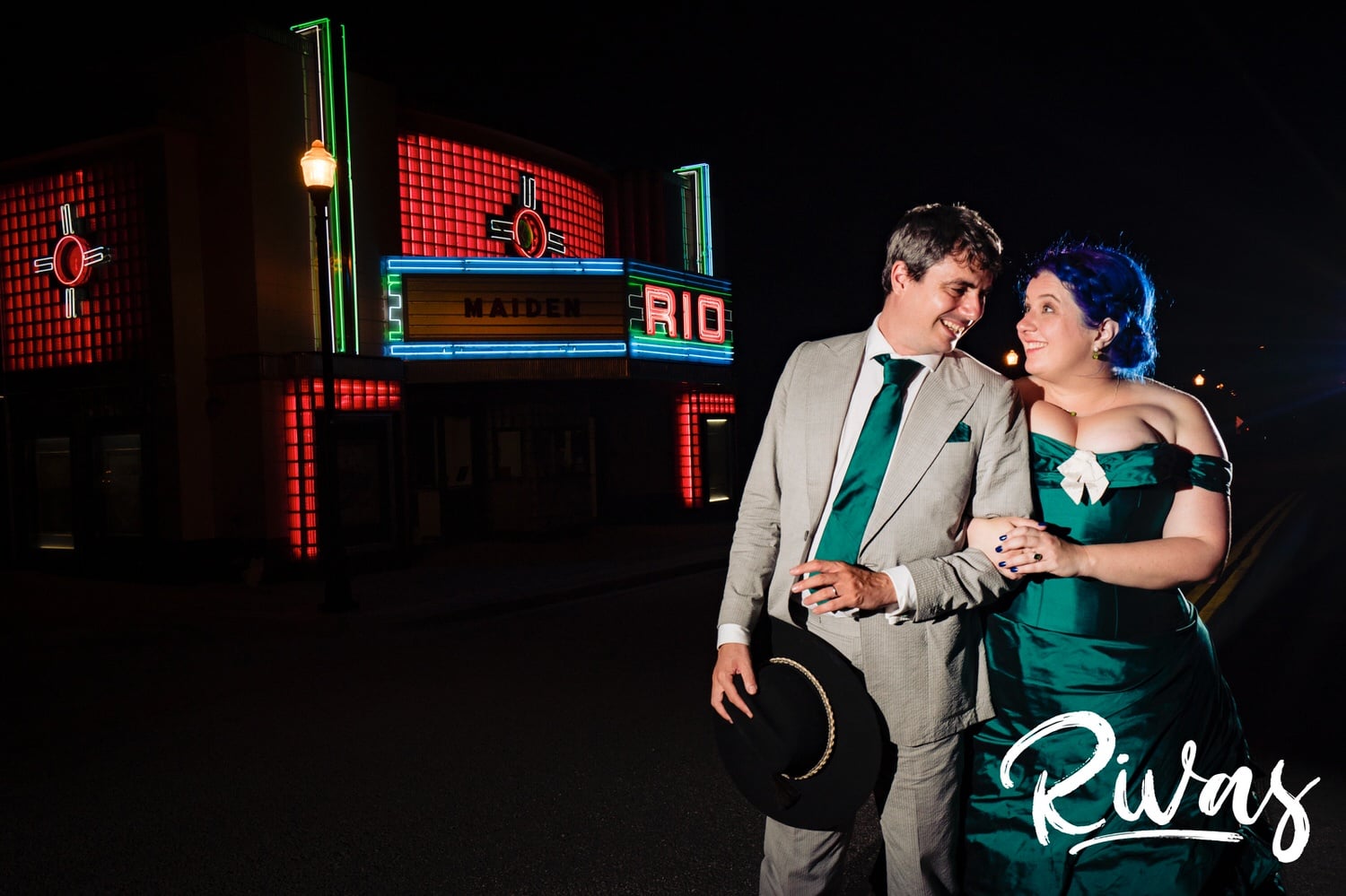 A vibrant portrait of a bride in a jade gown and a groom in cowboy boots standing shoulder to shoulder in front of the Rio Theater in downtown Overland Park just after dark on the day of their intimate backyard wedding. 