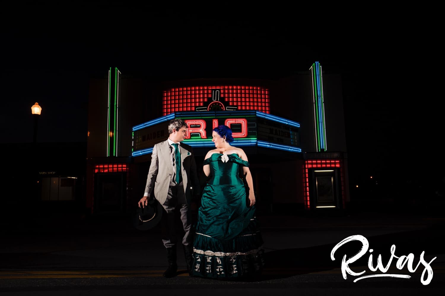 A vibrant portrait of a bride in a jade gown and a groom in cowboy boots standing shoulder to shoulder in front of the Rio Theater in downtown Overland Park just after dark on the day of their intimate backyard wedding. 