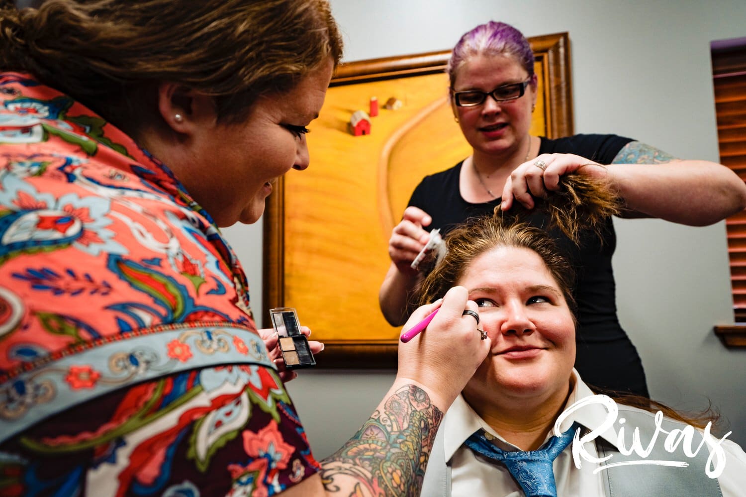 A colorful picture of a make-up artist leaning in to apply eye-liner to a woman in a tuxedo as a hair dresser straightens her hair on the morning of her wedding at Fire Stables in Kansas City. 