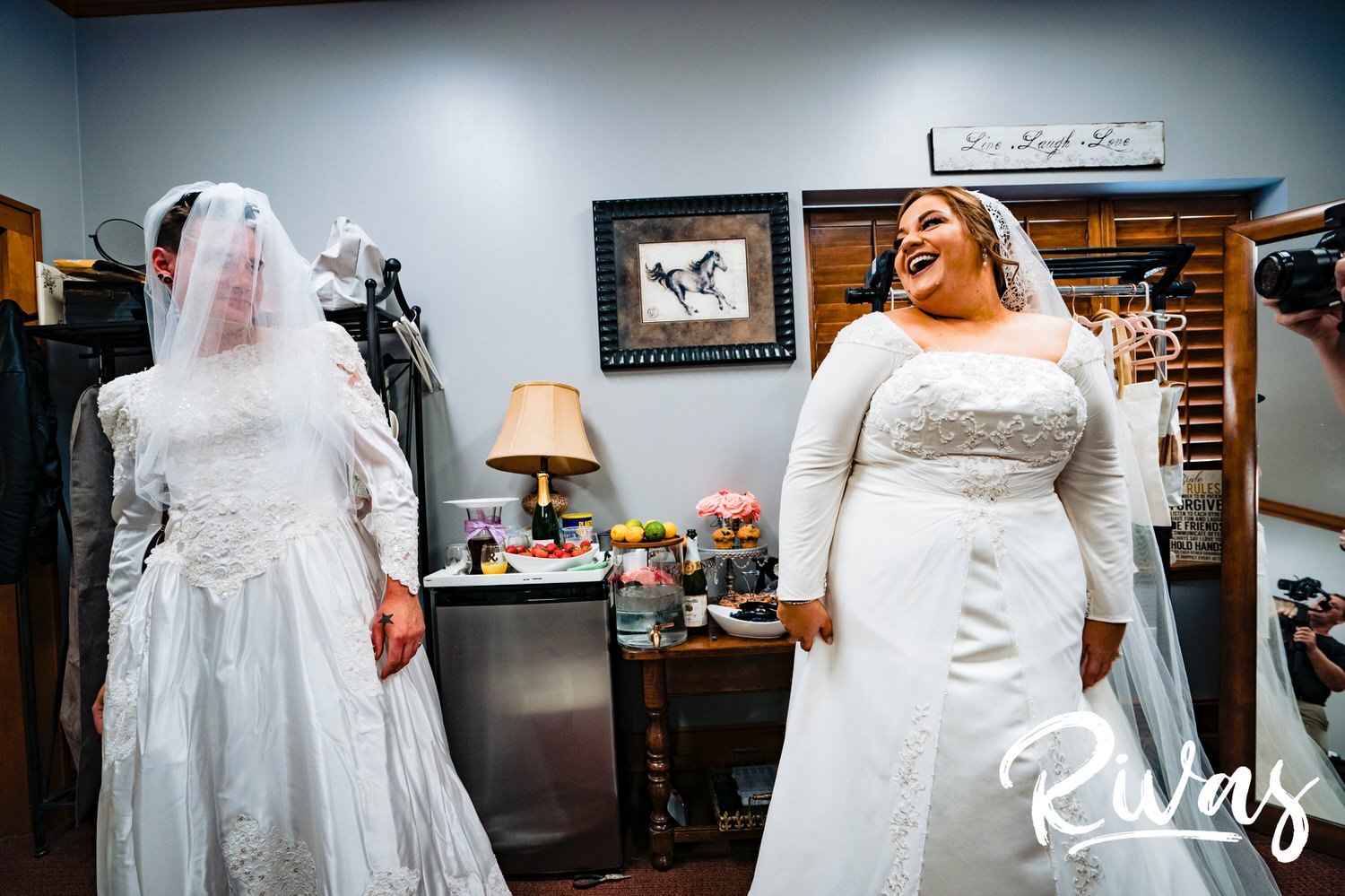 A candid picture of a bride and her brother-in-law, both in wedding dresses, laughing and looking at each other on the morning of a fire stables wedding. 