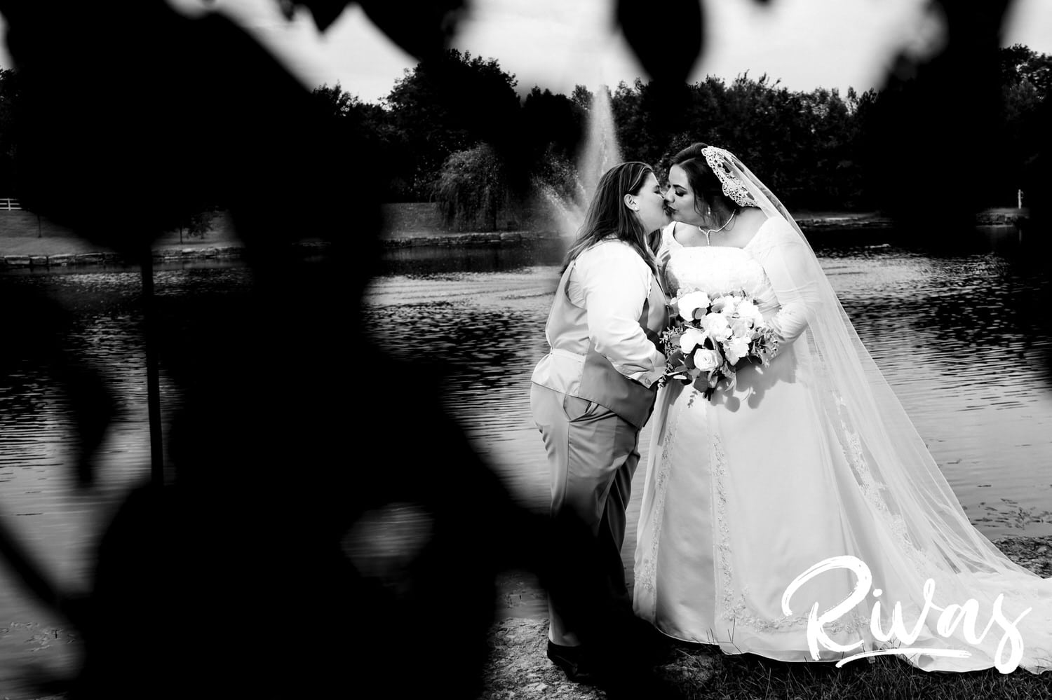 An intimate black and white portrait taken through the grass of two brides leaning in to share a kiss on their wedding day at Fire Stables. 