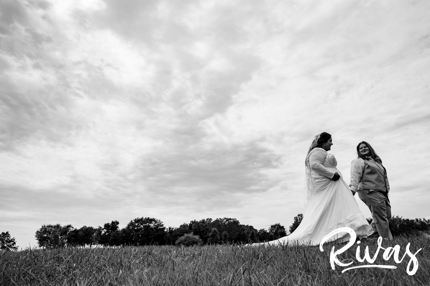 A candid black and white picture of two brides holding hands and walking across a lawn on their romantic wedding day at Fire Stables. 