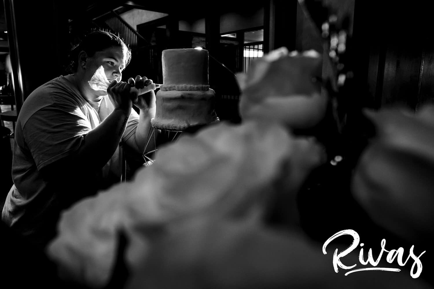 A candid black and white picture of a woman leaning in and adding a piped detail to a wedding cake on the morning of a wedding at Fire Stables. 