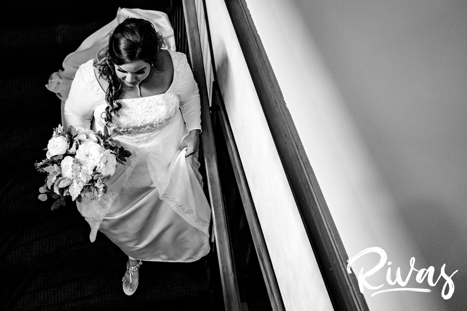 A candid black and white picture of a bride holding her bouquet and skirt as she walks down a flight of stairs to get married on her wedding day. 