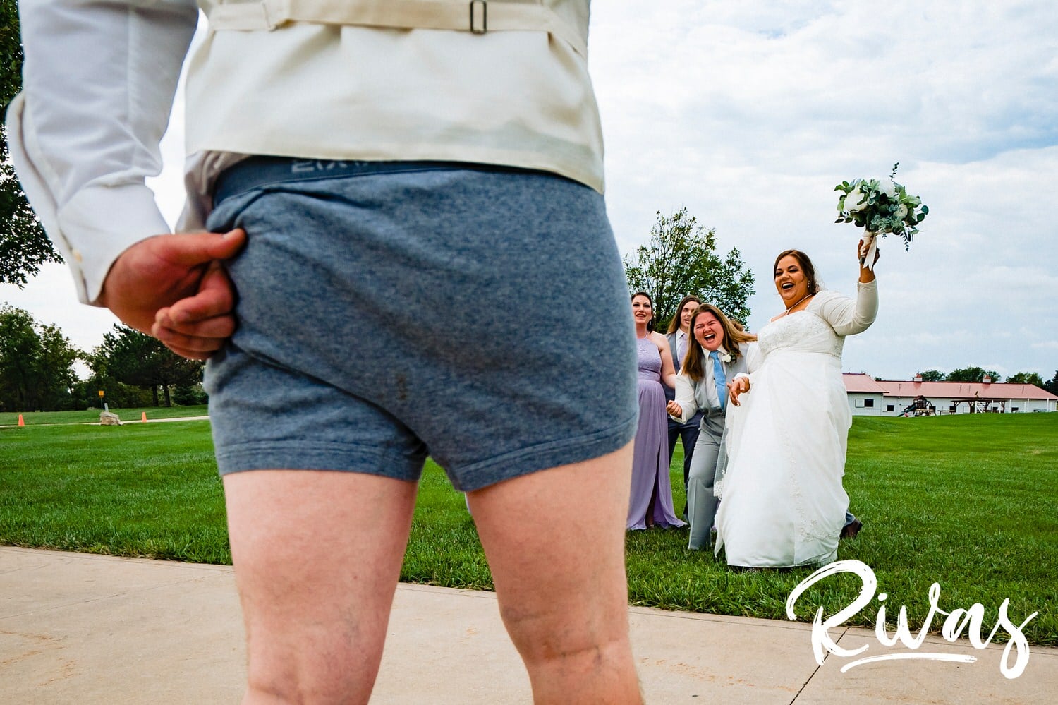 A candid picture of a man in half of a tuxedo with is pants around his ankles, boxer briefs sticking out, walking towards his sister who is laughing hysterically on her wedding day. 