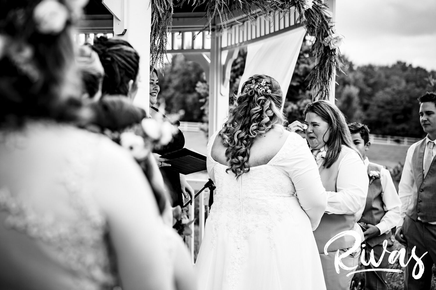 A candid black and white picture taken over the shoulders of bridesmaids of a bride wiping tears from her eyes during her wedding ceremony at Fire Stables. 