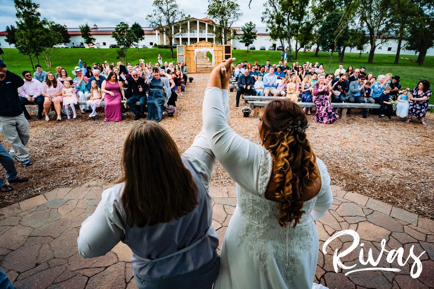 A candid, colorful picture taken from behind two brides raising their hands in celebration at the end of their wedding ceremony as their friends and family clap and celebrate. 