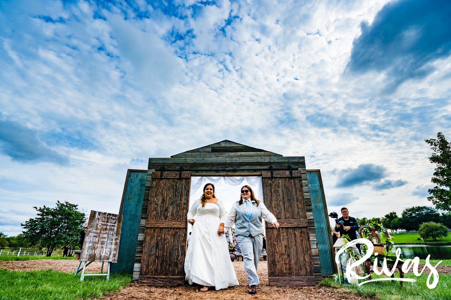 A candid, colorful picture of two brides holding hands and walking out of a set of rustic barn doors underneath a bright blue sky on their wedding day at Fire Stables. 