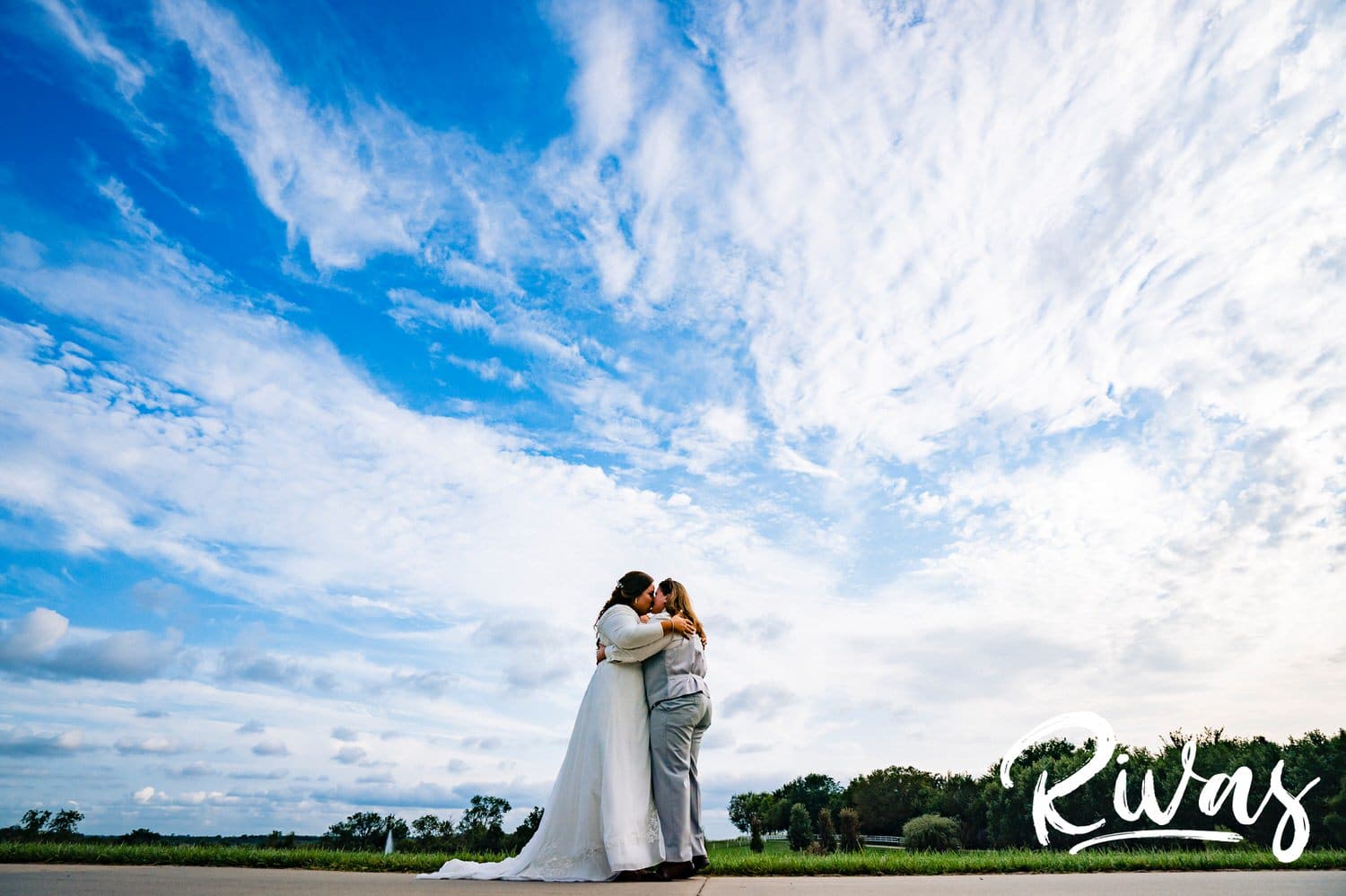 A colorful, wide picture of two brides sharing an embrace underneath a bright blue sky dotted with clouds on their wedding day at Fire Stables. 