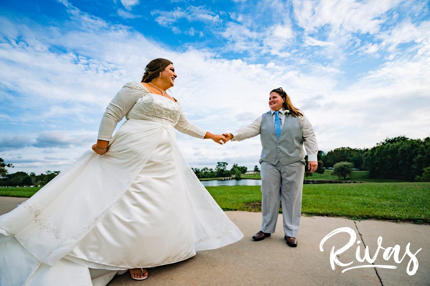 A colorful, wide picture of two brides sharing an embrace underneath a bright blue sky dotted with clouds on their wedding day at Fire Stables. 