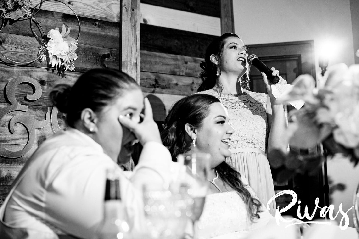 A candid black and white picture of a bride's sister giving a toast as a bride wipes tears from her eyes during a wedding reception at Fire Stables. 