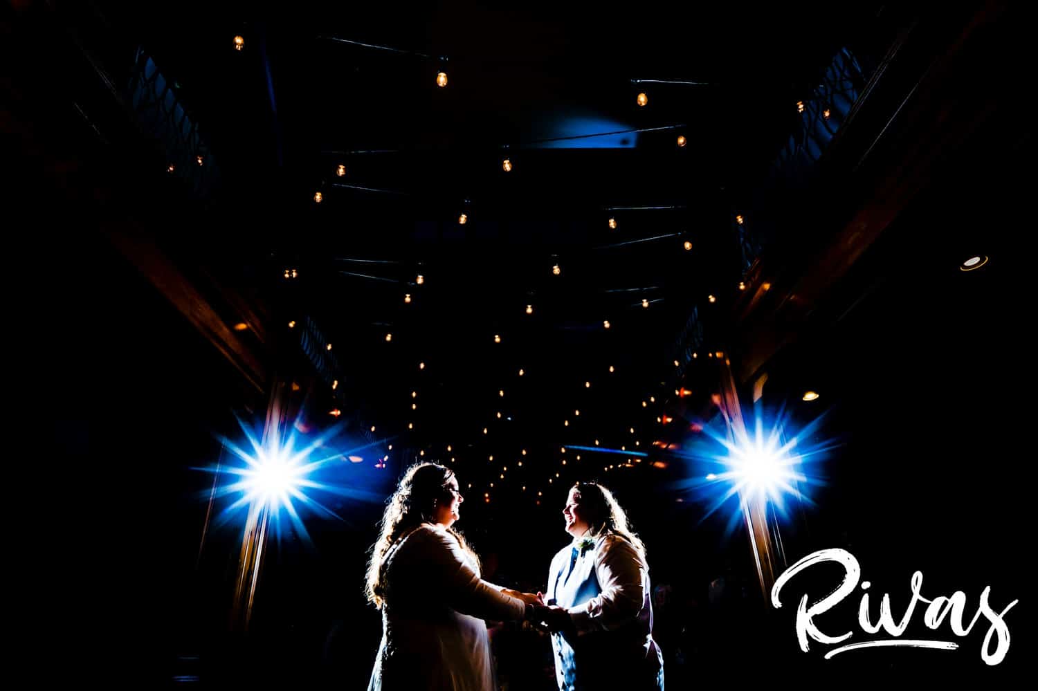 A colorful, wide picture of two brides holding hands as they dance together underneath a canopy of twinkly lights during their wedding reception at Fire Stables. 
