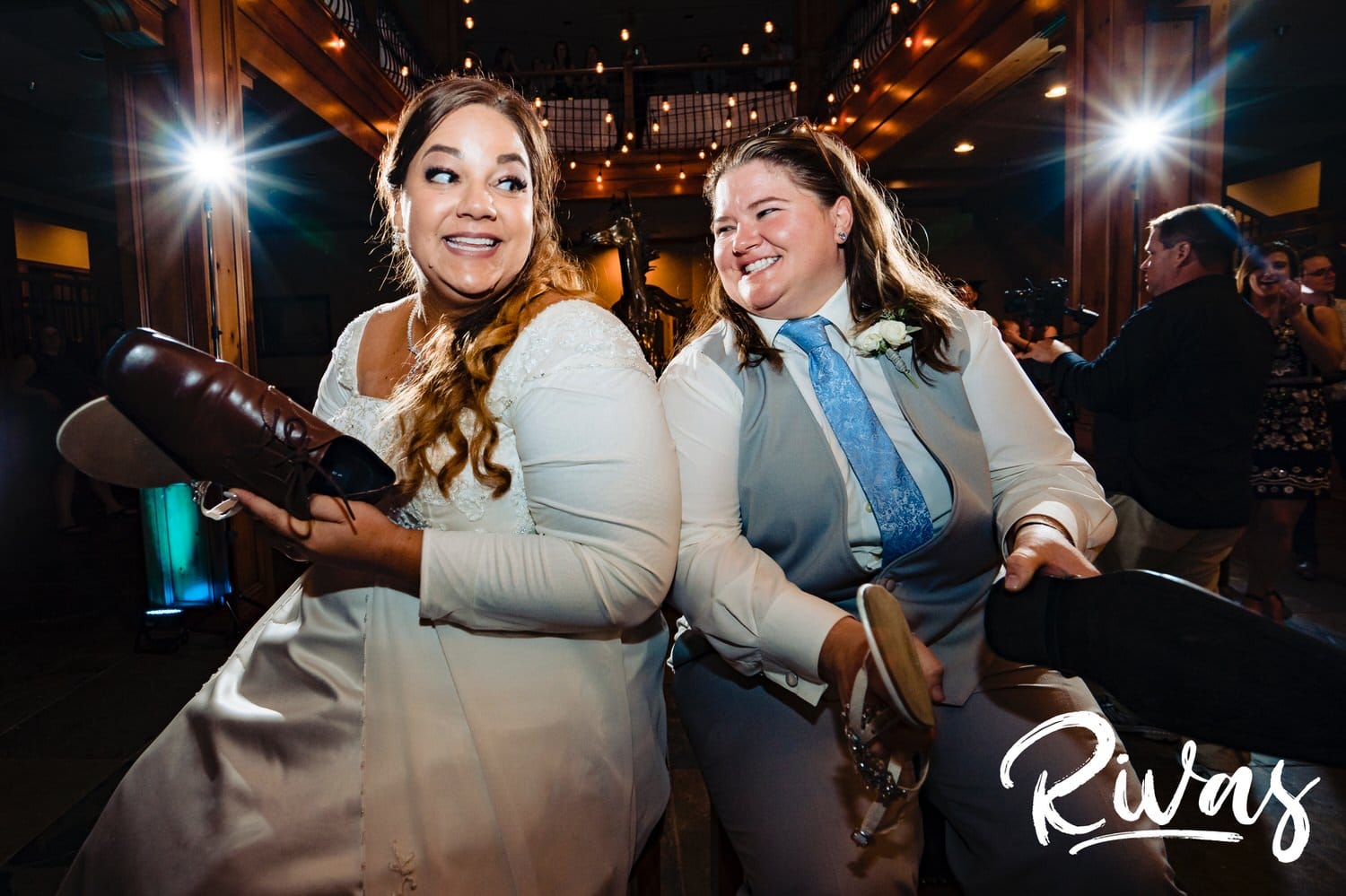 A candid picture of two brides, each holding one of their own shoes and one of the others' during the "shoe game" during their wedding reception at Fire Stables. 